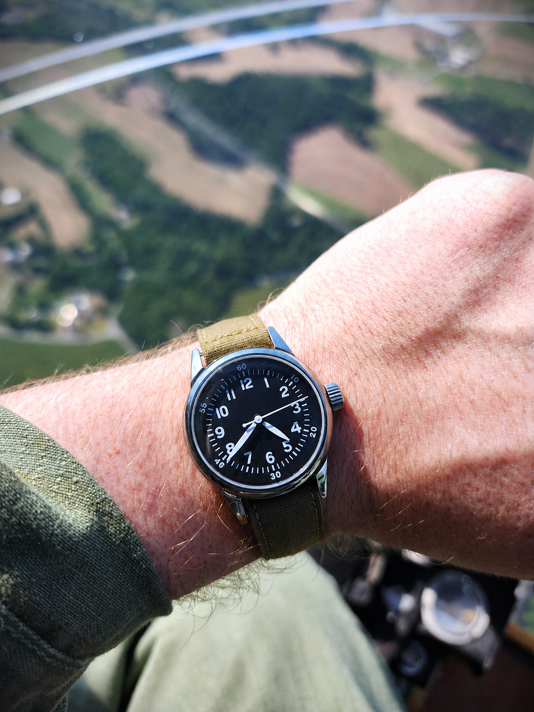 An A-11 with a tan strap and black dial worn on a person's wrist, photographed in an aircraft with a blurred landscape background visible through a window.