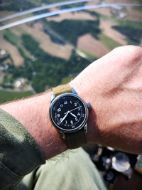 An A-11 with a tan strap and black dial worn on a person's wrist, photographed in an aircraft with a blurred landscape background visible through a window.