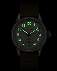 A WWII Ordnance Department wristwatch with a silver stainless steel case, an off-white dial with black Arabic numerals and hands, and an olive green canvas strap, shown in a low light environment to show the illuminated dial.