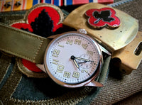 A WWII Ordnance Department wristwatch with a silver stainless steel case, an off-white dial with black Arabic numerals and hands, and an olive green canvas strap, shown against with WWII Medals and patches
