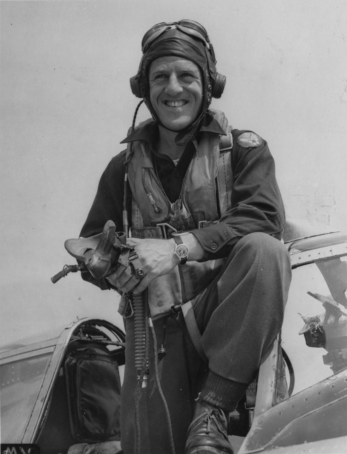 Black and white historical photograph of a smiling World War II fighter pilot wearing a flight helmet, headset, goggles, A-11 Wristwatch and a flight suit while sitting in the cockpit of an aircraft.