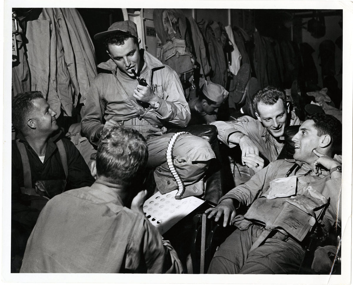 Black and white historical photograph showing a group of World War II soldiers laughing and smoking in a barracks or dressing area, with military uniforms and gear visible.