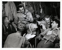 Black and white historical photograph showing a group of World War II soldiers laughing and smoking in a barracks or dressing area, with military uniforms and gear visible.