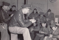 Black and white historical photograph showing a group of military personnel in uniform and leather jackets gathered in an indoor room, with one seated officer in the foreground hacking his watch with soldiers.